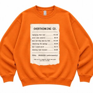 May include: A bright orange crewneck sweatshirt featuring a graphic print of a receipt titled OVERTHINKING CO. The receipt lists items like replaying that text, worst case scenario, and checking read receipts with prices, ending with TOTAL: PRICELESS (unfortunately).