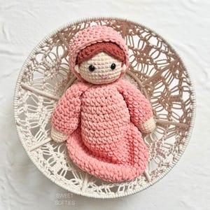 May include: A handmade crochet doll with a light pink hooded outfit, tan skin, and brown hair, featuring black safety eyes and a stitched mouth. The doll is positioned inside a round, off-white macrame basket. The text SWEET SOFTIES is visible.