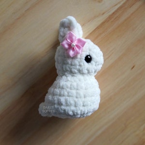 May include: White crocheted bunny with a pink flower on its head. The bunny has black eyes and a fluffy texture. The bunny is sitting on a wooden surface.