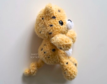 Puede incluir: Un pequeño juguete de peluche de guepardo con manchas negras sobre un fondo marrón claro. El juguete está de pie sobre sus patas traseras con las patas delanteras levantadas. El juguete tiene una barriga blanca y una mancha blanca en la cara.