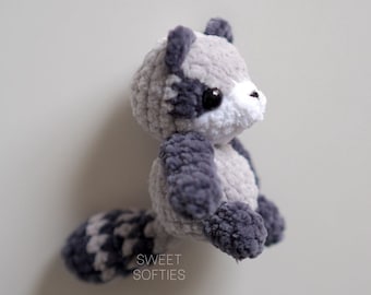 Puede incluir: Un peluche de mapache tejido a mano en tonos grises y blancos. El animal de peluche tiene detalles en gris oscuro en las orejas, patas y cola. El texto "SWEET SOFTIES" es visible. Un juguete suave y tierno.