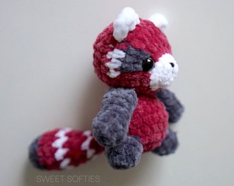 May include: A small crocheted red panda plush toy with white accents and grey arms. The toy has a red and white striped tail.