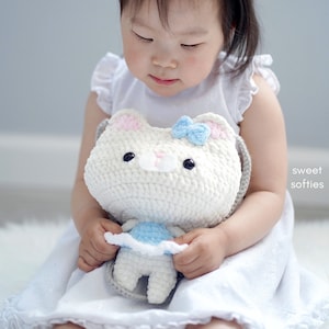 May include: A white crochet cat-shaped purse with a light blue bow and pink accents. The purse has black button eyes and a gray strap. A small, matching crochet doll is held inside. The image is labeled "sweet softies".