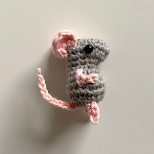 May include: A small grey crocheted mouse with pink ears and a pink tail. The mouse has black eyes and a stitched nose.
