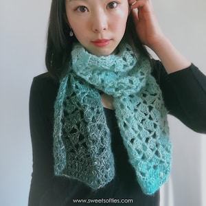 May include: A woman wearing a black shirt with a teal and green crochet scarf. The scarf has a gradient pattern and is crocheted with a delicate stitch.