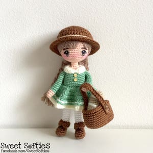 Könnte beinhalten: Eine gehäkelte Puppe mit einem braunen Hut, einem grünen Kleid und weißen Strumpfhosen. Die Puppe hält einen braunen Korb.