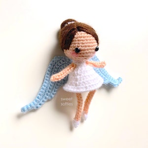 May include: A crocheted angel doll with brown hair, a white dress, and light blue wings. The doll is wearing white shoes and has black eyes.