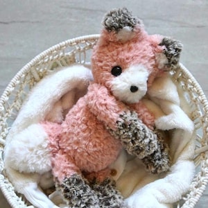 May include: A handmade plush fox toy in a white woven basket. The fox has pink fur with white and gray accents. It rests on a white blanket. The basket is round and the background is a light gray surface.