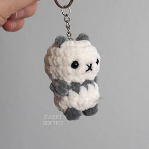 May include: White and gray crocheted panda bear keychain with black eyes. The keychain is attached to a silver key ring.