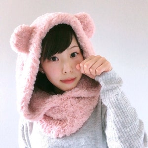 May include: A pink faux fur hooded scarf with bear ears. The scarf is worn by a person with dark hair and is styled to cover the neck and ears.