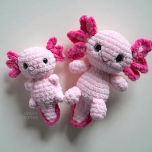 Könnte beinhalten: Zwei rosa gehäkelte Axolotl-Plüschtiere. Der größere Axolotl ist etwa 25 cm lang und der kleinere etwa 12 cm. Beide Axolotl haben rosa und weiße Wolle für ihre Kiemen und rosa Wolle für ihren Körper.