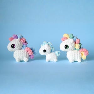 May include: Three white crochet unicorns with rainbow manes and tails. The unicorns have large, dark eyes and are displayed on a light blue backdrop. The text "SWEET SOFTIES" is visible in the bottom right corner.