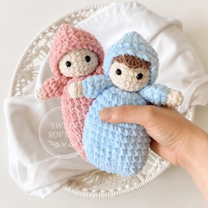 May include: Two handmade crochet dolls with soft yarn textures, one in light pink and one in light blue. Each doll features a hooded outfit, a cream-coloured face with black bead eyes, and brown hair. A circular logo with the text SWEET SOFTIES is visible.