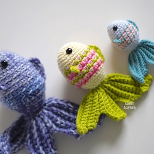 Puede incluir: Tres peces de juguete de ganchillo hechos a mano en tonos de azul, verde y crema. Los peces tienen ojos negros y aletas detalladas. Los peces est&aacute;n dispuestos sobre una superficie blanca. El texto "SWEET SOFTIES" es visible en la parte inferior derecha.