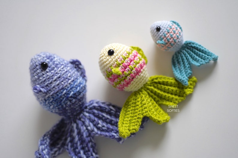 Puede incluir: Tres peces de juguete de ganchillo hechos a mano en tonos de azul, verde y crema. Los peces tienen ojos negros y aletas detalladas. Los peces est&aacute;n dispuestos sobre una superficie blanca. El texto "SWEET SOFTIES" es visible en la parte inferior derecha.