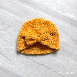 May include: A mustard yellow crocheted beanie with a large bow on top. The beanie is made of a soft, fuzzy yarn.