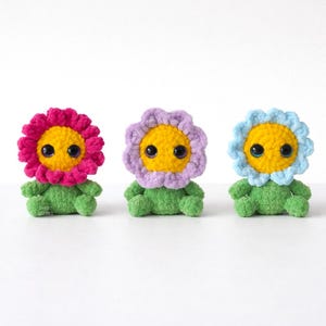May include: Three handmade crochet flower dolls. Each doll has a yellow center, a different colored petal (pink, purple, and blue), green body, and black eyes. The dolls are sitting upright.