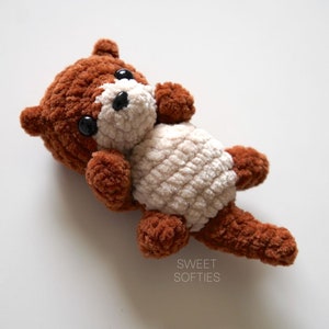 May include: A brown and white crocheted stuffed animal otter with black eyes. The otter is lying on its back with its legs in the air. The text "SWEET SOFTIES" is visible in the lower right corner of the image.