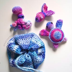 May include: A collection of handmade crochet toys, including an octopus, a fish, and a turtle, in shades of blue, purple, and pink. A matching drawstring bag is also included. The toys are arranged on a white surface.