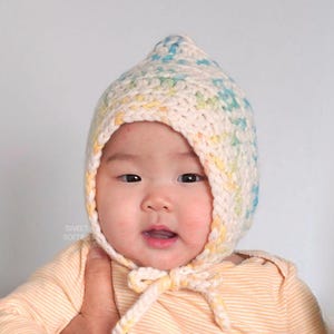 Könnte beinhalten: Ein Baby trägt eine gehäkelte Mütze mit einem gelben, blauen und weißen Ombre-Muster. Die Mütze hat einen Knoten am unteren Ende.