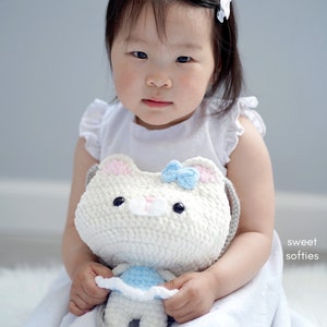 May include: A young child in a white dress holds a white crochet cat purse with a blue bow. The purse has black eyes and pink accents. The child is sitting on a white surface.