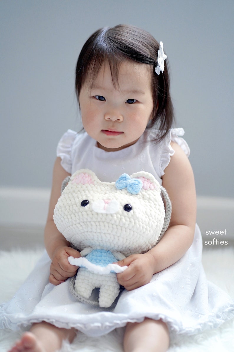 May include: A young child in a white dress holds a white crochet cat purse with a blue bow. The purse has black eyes and pink accents. The child is sitting on a white surface.