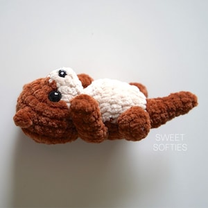 May include: A brown and cream crocheted stuffed otter toy with black eyes. The toy is lying on its back with its head tilted up.