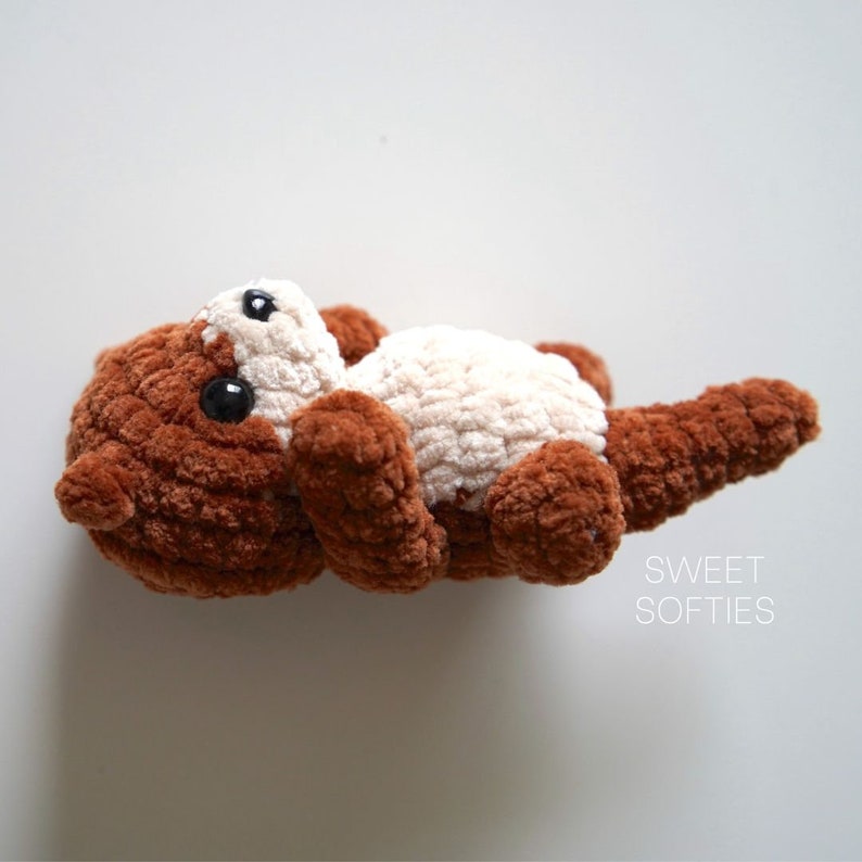 May include: A brown and cream crocheted stuffed otter toy with black eyes. The toy is lying on its back with its head tilted up.