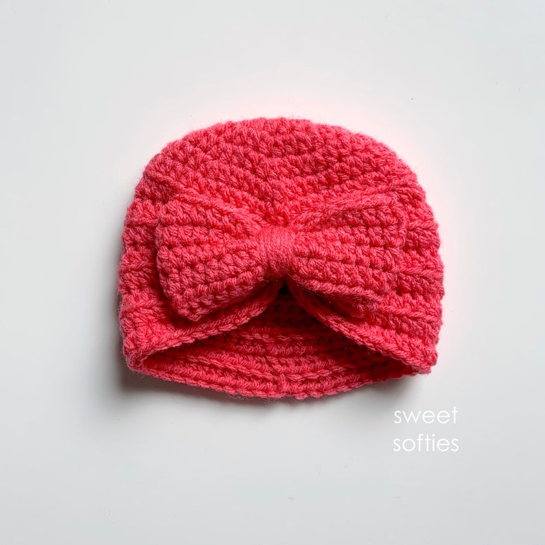 May include: A pink crocheted turban with a large bow on top. The turban is made with a soft, textured yarn.