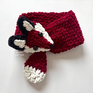 May include: A handmade, plush fox scarf in burgundy, white, and black. The scarf features a fox head with button eyes and a tail. The text "SWEET SOFTIES" is visible on the scarf.