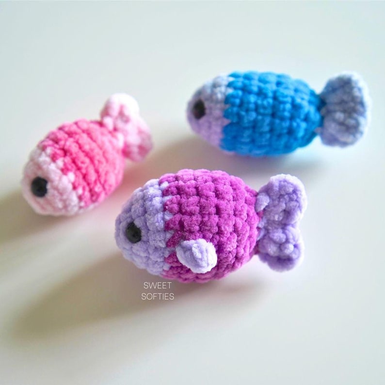 Op de afbeelding: Drie gehaakte vis speelgoed in roze, paars en blauw. De vissen zijn gemaakt met een pluche garen en hebben zwarte ogen.