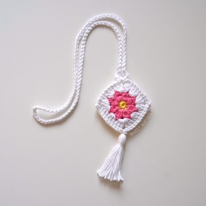 May include: A white crochet necklace with a square pendant featuring a pink and yellow flower design. The pendant has a white border and a white tassel. The necklace is handmade and has a braided cord.