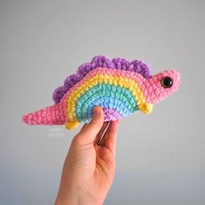 May include: A rainbow colored crocheted dinosaur toy with large black eyes. The dinosaur is pink, yellow, green, blue, and purple.