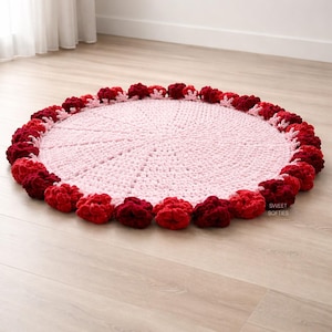 May include: A round, handmade crochet rug in soft pink with a border of red and burgundy pom-poms. The rug is placed on a light wood floor. The text "Sweet Softies" is visible on the rug.