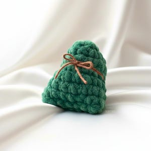 May include: A handmade, green crocheted item, possibly a decorative piece or toy, tied with a brown string bow. The item has a textured, bumpy surface and is set against a white, silky background.