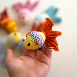 Puede incluir: Un pez dorado de ganchillo con un dise&ntilde;o amarillo, arco&iacute;ris y naranja. El pez tiene un ojo negro y una aleta caudal naranja. Otros peces de ganchillo de varios colores est&aacute;n en el fondo. El texto "SWEET SOFTIES" est&aacute; en la esquina inferior derecha.
