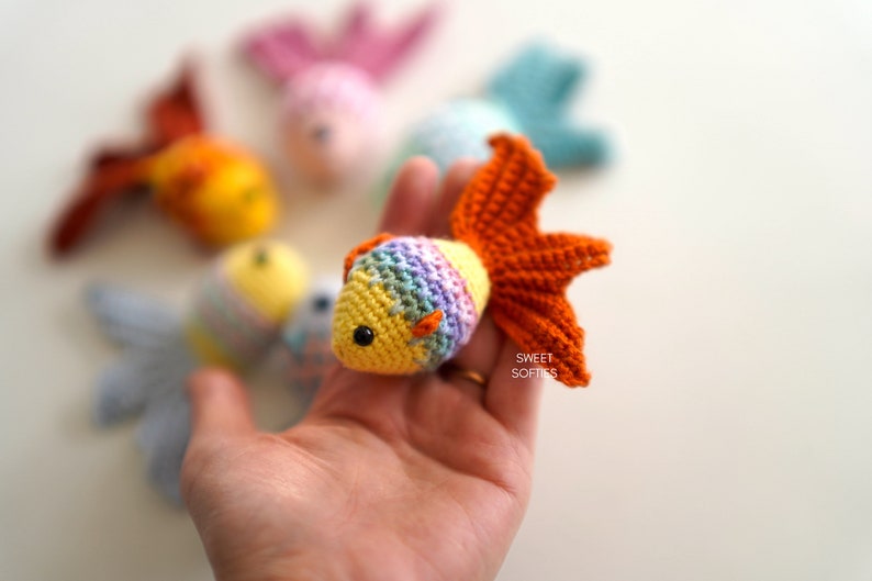 Puede incluir: Un pez dorado de ganchillo con un dise&ntilde;o amarillo, arco&iacute;ris y naranja. El pez tiene un ojo negro y una aleta caudal naranja. Otros peces de ganchillo de varios colores est&aacute;n en el fondo. El texto "SWEET SOFTIES" est&aacute; en la esquina inferior derecha.