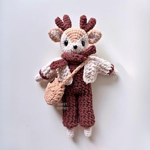 May include: A handmade crochet deer doll wearing a burgundy jumpsuit, a cream-colored jacket, and a matching scarf. The deer has brown antlers, a small tan satchel, and the text "SWEET SOFTIES" on the side. The doll is approximately 10 inches tall.