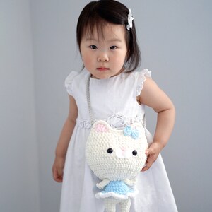 May include: A white crochet cat-shaped crossbody bag with a blue bow and dress, held by a child wearing a white dress. The bag has black button eyes and pink accents on the ears. The child is wearing a white hair clip.