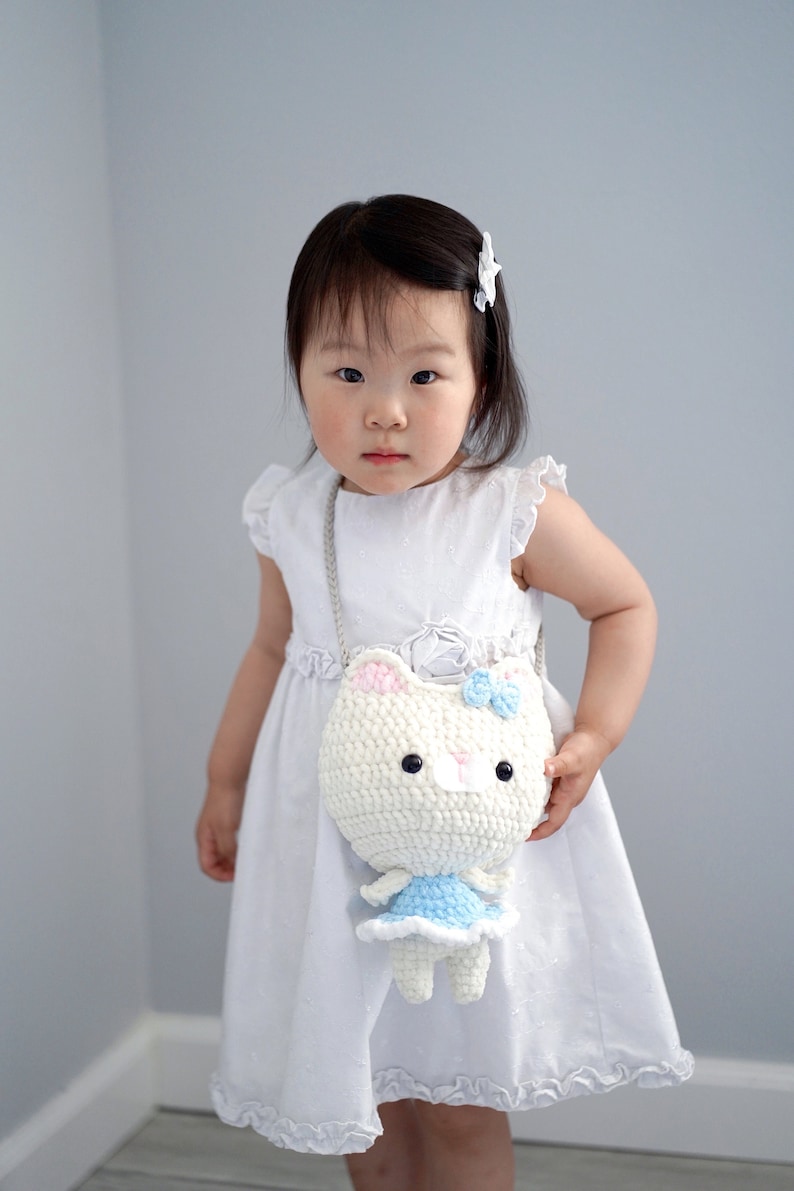 May include: A white crochet cat-shaped crossbody bag with a blue bow and dress, held by a child wearing a white dress. The bag has black button eyes and pink accents on the ears. The child is wearing a white hair clip.