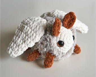 May include: A crocheted plush toy resembling a moth with white wings, a brown fuzzy body, and large brown eyes. The toy is sitting on a white surface. The text "SWEET SOFTIES" is visible on the toy.