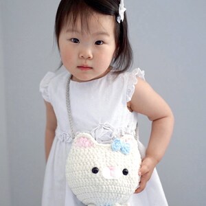 May include: A white crochet cat purse with pink ears and a blue bow, held by a child wearing a white dress. The purse has a long silver chain strap. The text "sweet softies" is in the bottom left corner.