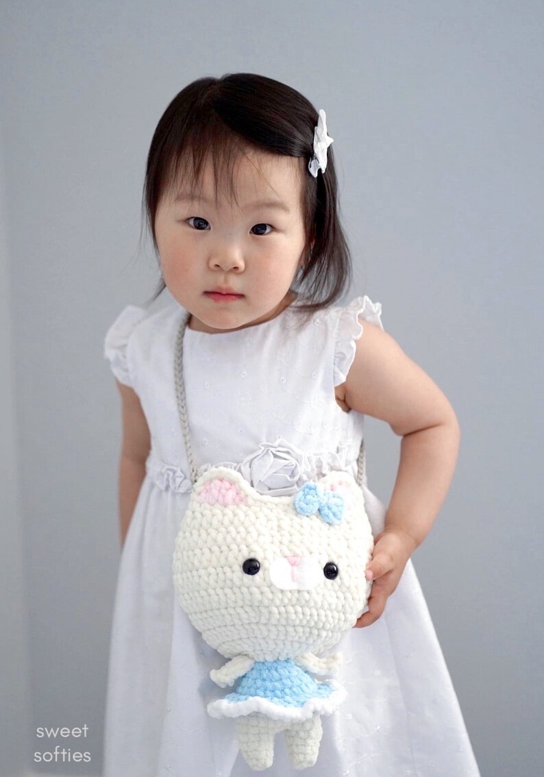 May include: A white crochet cat purse with pink ears and a blue bow, held by a child wearing a white dress. The purse has a long silver chain strap. The text "sweet softies" is in the bottom left corner.