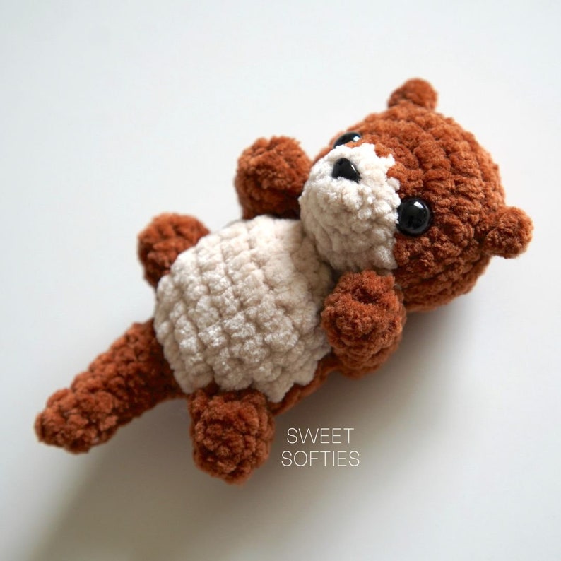 May include: A brown and cream crocheted stuffed otter toy with black eyes. The toy is lying on its back with its legs in the air. The text "SWEET SOFTIES" is printed below the toy.