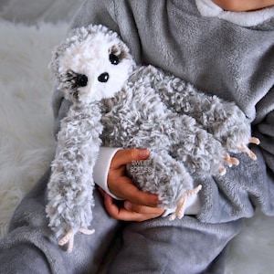 Puede incluir: Un peluche de perezoso gris con cara blanca y ojos negros es sostenido en brazos. El perezoso tiene pelo largo y suave y pequeñas garras beige. El texto "SWEET SOFTIES" es visible en la manga de la persona que sostiene el juguete.