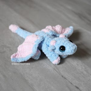 以下が含まれることがあります： 水色とピンクの柔らかい手作りのぬいぐるみドラゴン。ドラゴンは大きくて丸い目と詳細な翼を持っています。ぬいぐるみはふわふわした素材でできており、灰色の表面に置かれています。「@SWEETSOFTIES」の文字が見えます。