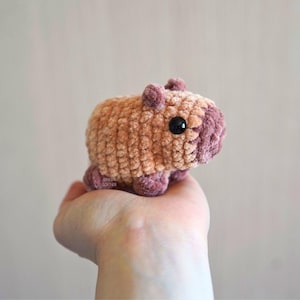 Puede incluir: Un juguete de capibara de peluche hecho a mano, en tonos marrones y malvas. El animal de ganchillo tiene un cuerpo redondeado, orejas pequeñas y ojos negros. El juguete es suave y tierno, ideal para niños o como objeto decorativo.