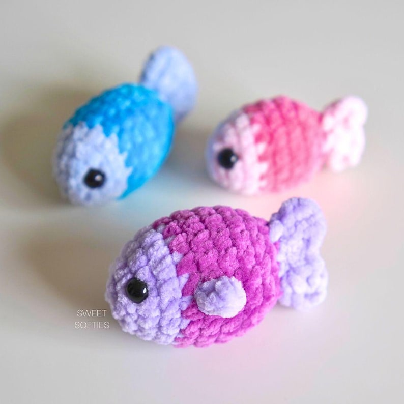 Op de afbeelding: Drie gehaakte vis speelgoed in verschillende kleuren: blauw, roze en paars. De vissen hebben zwarte ogen en zijn gemaakt van zacht, pluche garen.