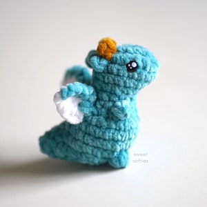 May include: A teal blue crocheted dragon with white wings and a yellow horn. The dragon has black eyes and a sweet expression.