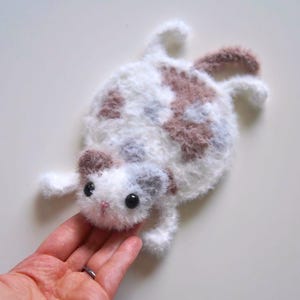May include: A white and brown crocheted cat toy with black eyes. The cat is lying on its back with its paws in the air.